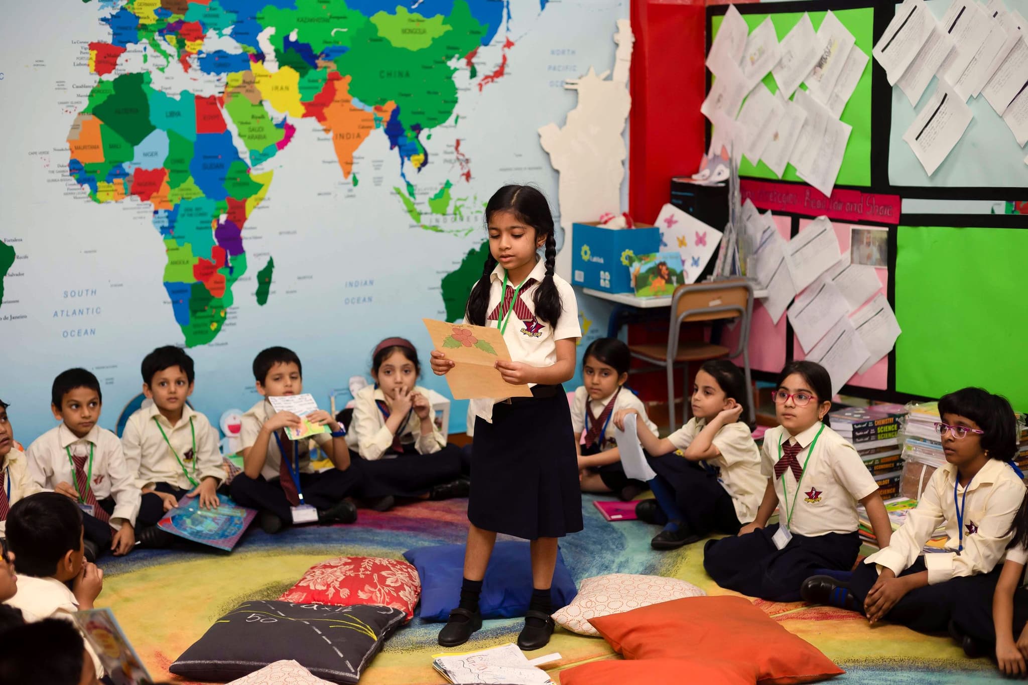 A primary pupil reads her work aloud to classmates seated on cushions beneath a world map