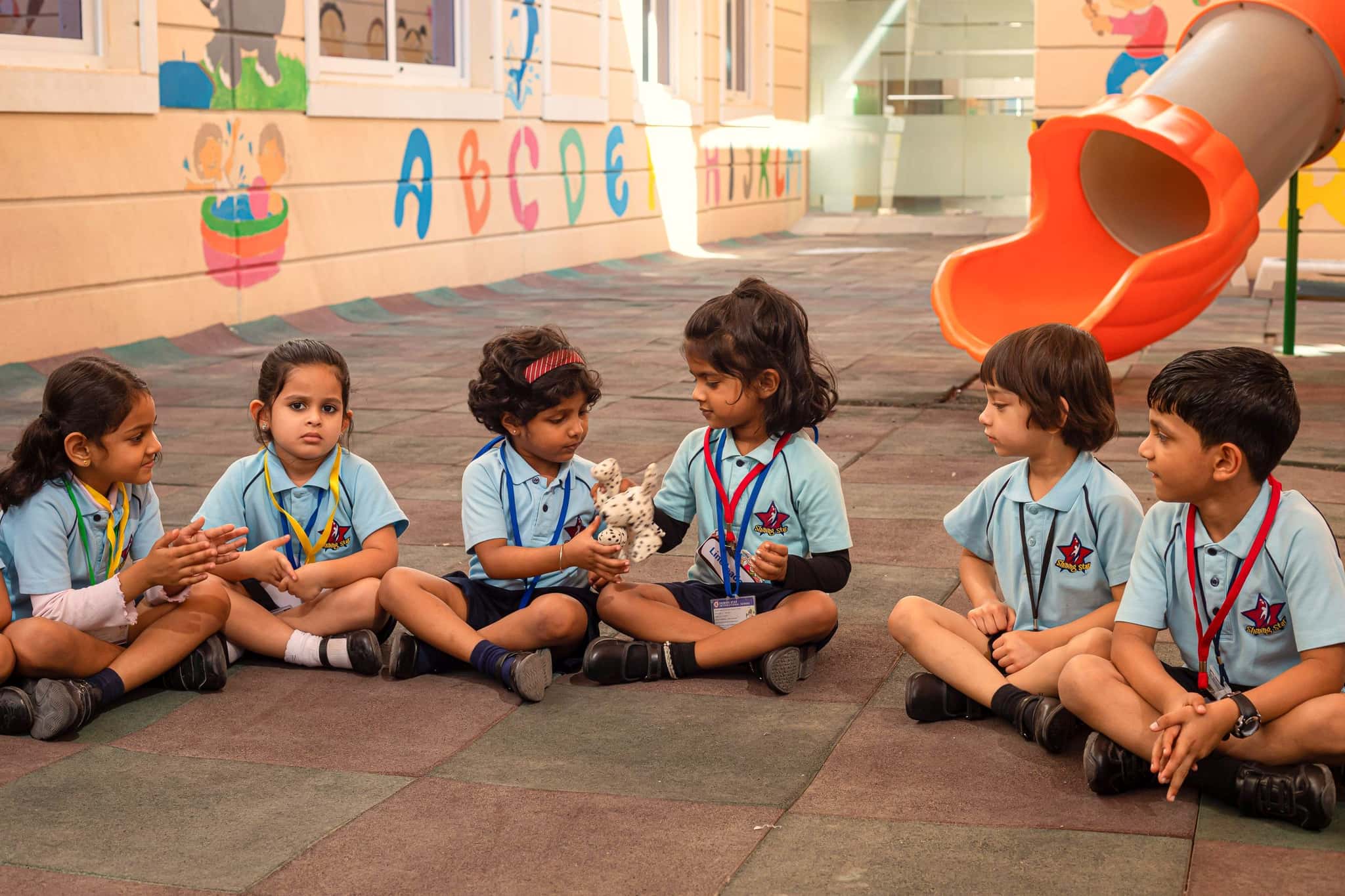 Kindergarten pupils sitting in a circle on the shaded play court, one girl holding up a soft toy for the group