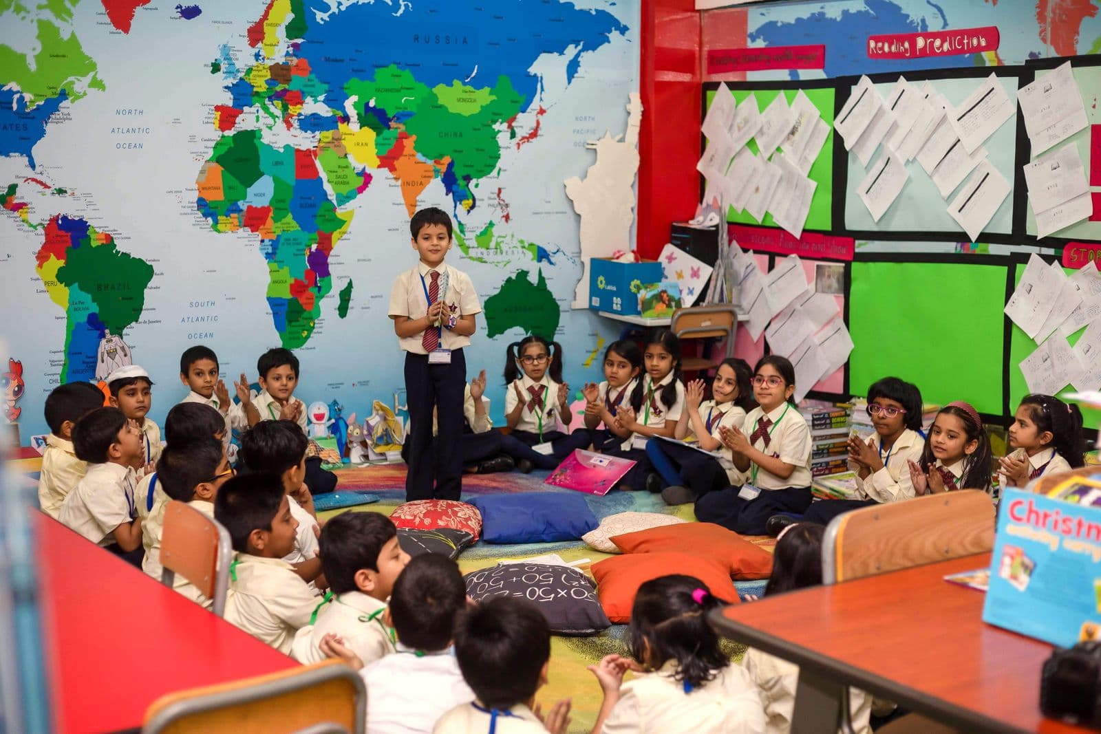 A primary pupil stands to present while classmates listen from floor cushions