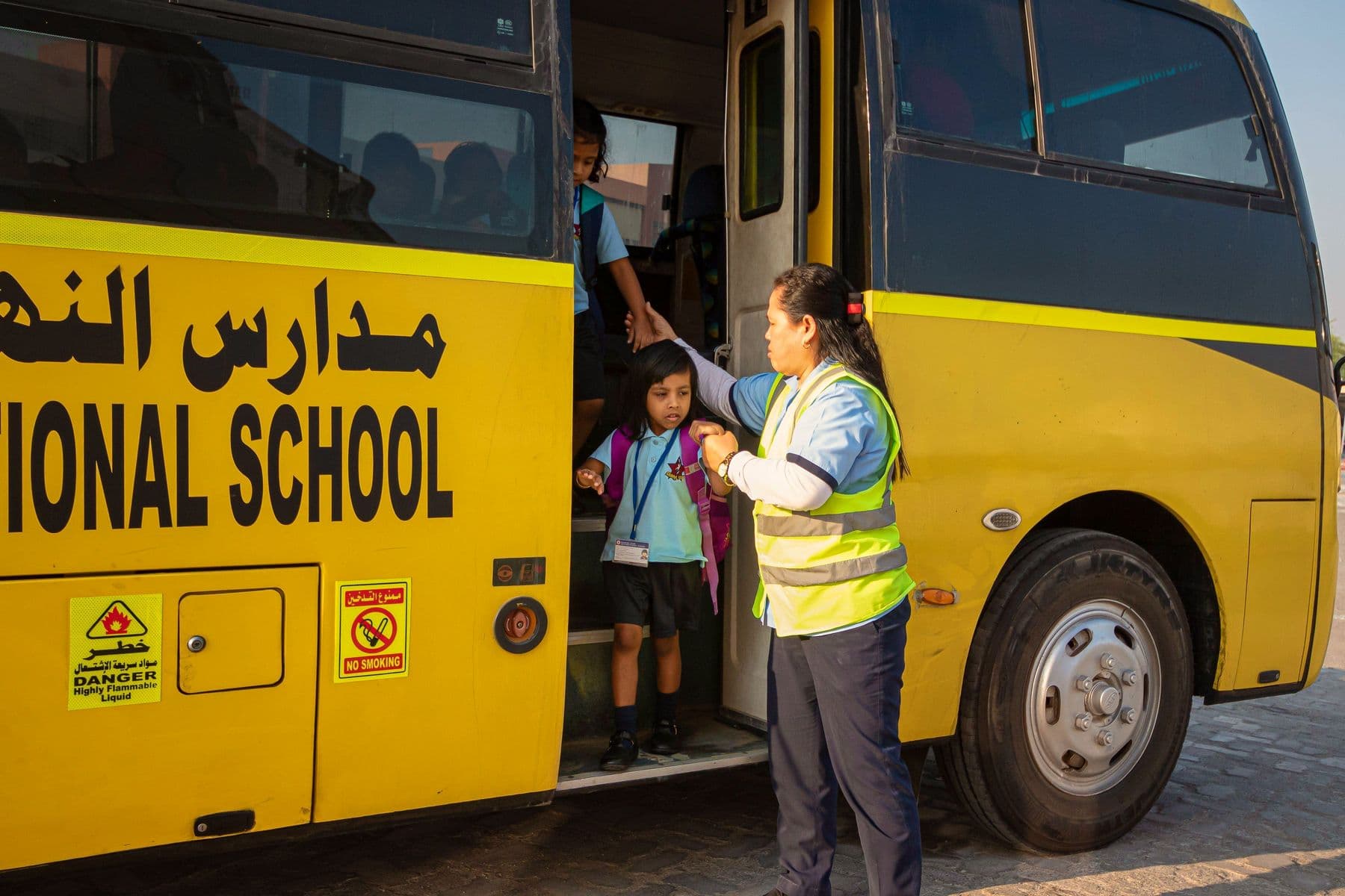 A bus attendant in a high-visibility vest helps a young pupil down the steps of a yellow school bus